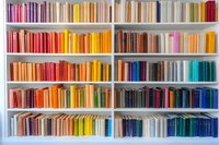 Books arranged by color on white wooden shelves creating rainbow spectrum pattern. Organized bookshelf displays harmonious gradient from pink through yellow to blue tones.