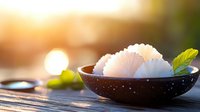 White rice balls in black ceramic bowl with green leaf garnish on wooden surface at sunset, soft bokeh background creates warm atmospheric dining scene.