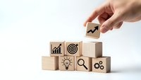 Hand placing wooden cube with growth arrow onto stack of business icon blocks representing strategy development and goal achievement.