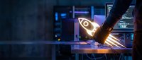 Welder in protective gear creating sparks while joining metal parts in industrial workshop with glowing neon rocket symbol on workbench.