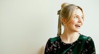 Young woman with blonde hair styled in a bun, fair complexion, wearing a dark green floral top, smiling and looking to the side against a neutral white background for portrait and lifestyle use.