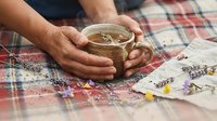 Hands holding warm herbal tea cup with lavender flowers on cozy plaid blanket for relaxation and wellness moments.