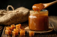 Golden caramel sauce dripping from wooden spoon into glass jar with caramel candies and burlap sack on rustic wooden background for dessert recipes.