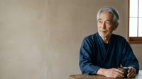 Senior Asian man with gray hair wearing dark blue casual shirt, seated indoors with neutral beige wall background and natural window light, contemplative portrait for lifestyle and editorial use.