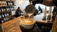 Barista pouring steamed milk creating latte art in coffee cup at modern cafe with customers in background. Professional coffee making process in cozy coffeehouse atmosphere.