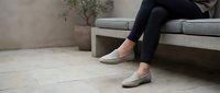 Woman in black leggings wearing beige ballet flats sitting on modern gray sofa in minimalist living room with potted plant and neutral decor.