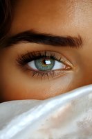 Close-up of bright green eye with long eyelashes and defined eyebrow, partially covered by white fabric, showing mystery and allure on tanned skin tone.