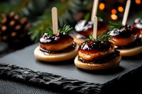 Mini foie gras appetizers on round crackers garnished with cranberries and pine sprigs, served on black slate plate with bokeh lights in background.