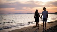 Young couple walking hand in hand on a sandy beach at sunset, silhouetted against a vibrant pink and purple sky over calm ocean waters.