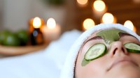 Woman relaxing with cucumber slices on eyes and green facial mask during spa treatment with candles creating peaceful wellness atmosphere.