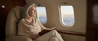 Elegant blonde businesswoman reading book in luxury private jet cabin during flight with scenic sky view through windows for corporate travel concepts.