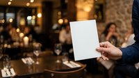Man in formal navy suit holding blank white card at elegant dinner event with warm ambient lighting and blurred guests in background.