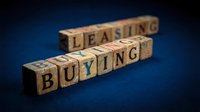 Wooden blocks spelling leasing and buying on dark blue background, representing real estate investment decisions and property acquisition options.