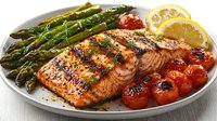 Grilled salmon fillet served with roasted asparagus, cherry tomatoes and lemon slices on white plate, garnished with fresh herbs and black pepper.