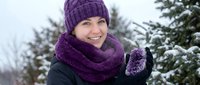 Happy young Caucasian woman in purple knit hat and scarf smiling outdoors in snowy winter landscape with evergreen trees for seasonal lifestyle content.