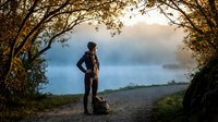Young woman hiker standing on mountain trail overlooking misty lake at sunrise, wearing backpack and outdoor gear, peaceful morning adventure scene.