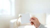 Hand holding blank white square gift tag with twine string against bright window light and neutral interior background for product labeling and branding purposes.