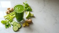 Fresh green smoothie with celery, cucumber, ginger and herbs on marble counter. Healthy detox drink surrounded by organic vegetables for wellness lifestyle.