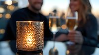 Romantic evening ambiance with glowing candlelight in textured glass holder, champagne flutes and blurred couple in background creating intimate dining atmosphere.