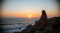 Young woman meditating in lotus position on rocky coastline during golden sunset over ocean waves, peaceful mindfulness practice by the sea.
