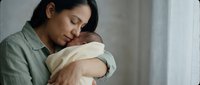 Tender moment of Asian mother gently holding her sleeping newborn baby in soft natural light, expressing pure maternal love and bonding in peaceful home setting.