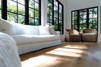 Modern bright sunroom with white sofa, rattan chairs, and large black-framed windows overlooking lush garden. Natural light streams across hardwood flooring creating warm ambiance.