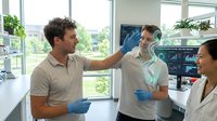 Young Caucasian male doctor examining patient with stethoscope in modern medical office while Asian female colleague observes professional healthcare consultation.