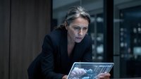 Middle-aged woman with gray hair and glasses wearing black blazer, focused on laptop screen in modern office environment with blurred background lighting.