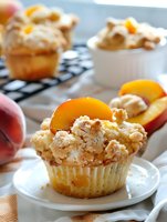 Fresh peach streusel muffin topped with crumbly cinnamon sugar topping and sliced ripe peaches on white plate, selective focus on golden pastry texture.