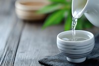 Pouring hot water from white ceramic teapot into traditional tea cup on dark wooden surface with green leaves creating peaceful zen atmosphere.