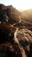 Winding mountain road cutting through dramatic rocky terrain with golden sunlight illuminating arid landscape, perfect for travel and adventure design projects.