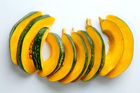 Fresh sliced kabocha pumpkin arranged in curved pattern on white background showing bright yellow flesh and dark green skin, ready for cooking or presentation.