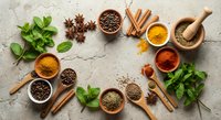 Aromatic spices and herbs arranged in wooden bowls with mortar and pestle on textured stone background for culinary cooking and seasoning recipes.