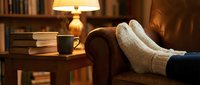 Cozy home relaxation scene with feet in warm wool socks resting on leather armchair, steaming mug and books on side table, warm lamp light.