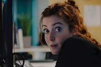 Young Caucasian woman with surprised expression looking at computer screen in office setting, messy bun hairstyle and casual attire, captured in candid moment.