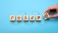 Hand placing wooden cube with target icon completing business strategy sequence on blue background for goal achievement and success planning.