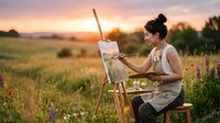 Female artist with dark hair in bun, wearing beige apron, painting wildflower meadow landscape at golden hour sunset. Outdoor art creation.