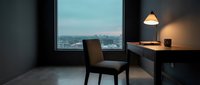 Modern office workspace with floor-to-ceiling window overlooking cityscape at dusk, minimalist desk setup with pendant lamp and chair for corporate or remote work environments.