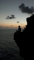 Silhouette of young man sitting on cliff edge overlooking ocean at sunset, dramatic dark clouds and peaceful seascape create contemplative atmosphere.