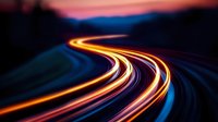 Long exposure light trails on winding mountain road at dusk create glowing orange and red streaks against dark blue twilight sky, capturing motion and speed.