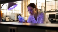 Young female scientist in white lab coat examining sample under purple UV light in modern laboratory setting for research and analysis work.