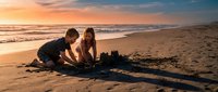 Young couple building sandcastle together on beach during golden hour sunset with ocean waves and romantic sky creating perfect vacation moment.