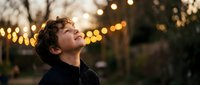 Young boy with curly hair looking up with wonder and joy during golden hour with warm bokeh lights creating magical atmosphere for family portraits.