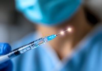 Medical professional in surgical mask and blue gloves holding syringe with glowing needle tip against blurred hospital background, dramatic lighting effect.