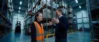 Professional warehouse managers discussing logistics operations in modern distribution center with high storage racks and industrial lighting.