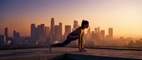 Athletic woman doing push-ups on urban rooftop at sunrise with city skyline backdrop, fitness workout exercise training outdoors in morning light.