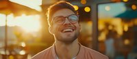 Young Caucasian man with glasses and beard wearing pink t-shirt laughing genuinely during golden hour sunset, bokeh lights in background create warm atmosphere.