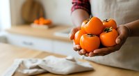 Woman holding fresh orange tomatoes in kitchen with wooden cutting board and blurred background, suitable for food, cooking, and healthy eating content.