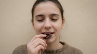 Young Caucasian woman enjoying dark chocolate piece with closed eyes and peaceful expression wearing beige sweater against neutral background.
