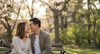 Young Asian couple sharing intimate moment on park bench during spring cherry blossom season with warm golden hour lighting and romantic atmosphere.